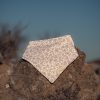 Bandana para bebé hecha a mano con estampado de pequeñas flores rosas sobre fondo claro, colocada sobre una piedra al aire libre.