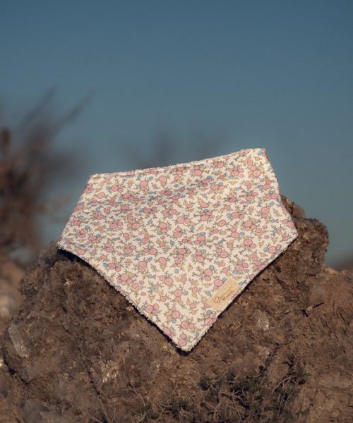 Bandana para bebé hecha a mano con estampado de pequeñas flores rosas sobre fondo claro, colocada sobre una piedra al aire libre.