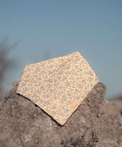 Bandana para bebé hecha a mano con estampado de pequeñas flores silvestres sobre fondo claro, colocada sobre una piedra al aire libre.