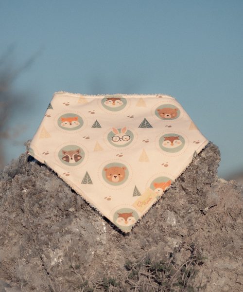 Bandana para bebé hecha a mano con estampado de animales del bosque sobre fondo beige, colocada sobre una piedra al aire libre.