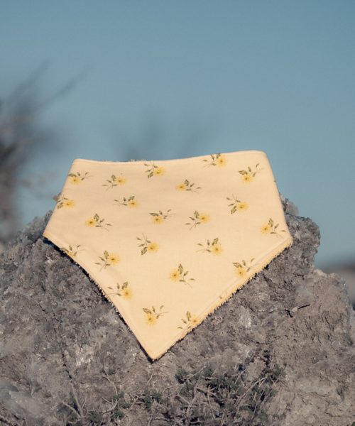 Bandana para bebé hecha a mano con estampado de pequeñas flores amarillas sobre fondo crema, colocada sobre una piedra al aire libre.